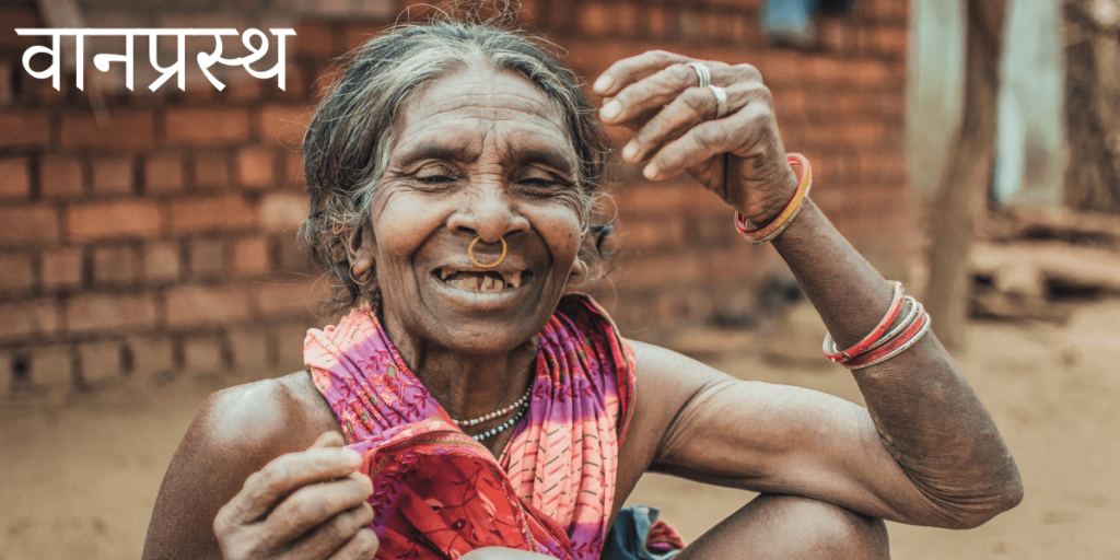 Une femme âgée souriante assise sur le bord de la route avec le titre en Sanskrit वानप्रस्थ – Vānaprasta – Retrait spirituel